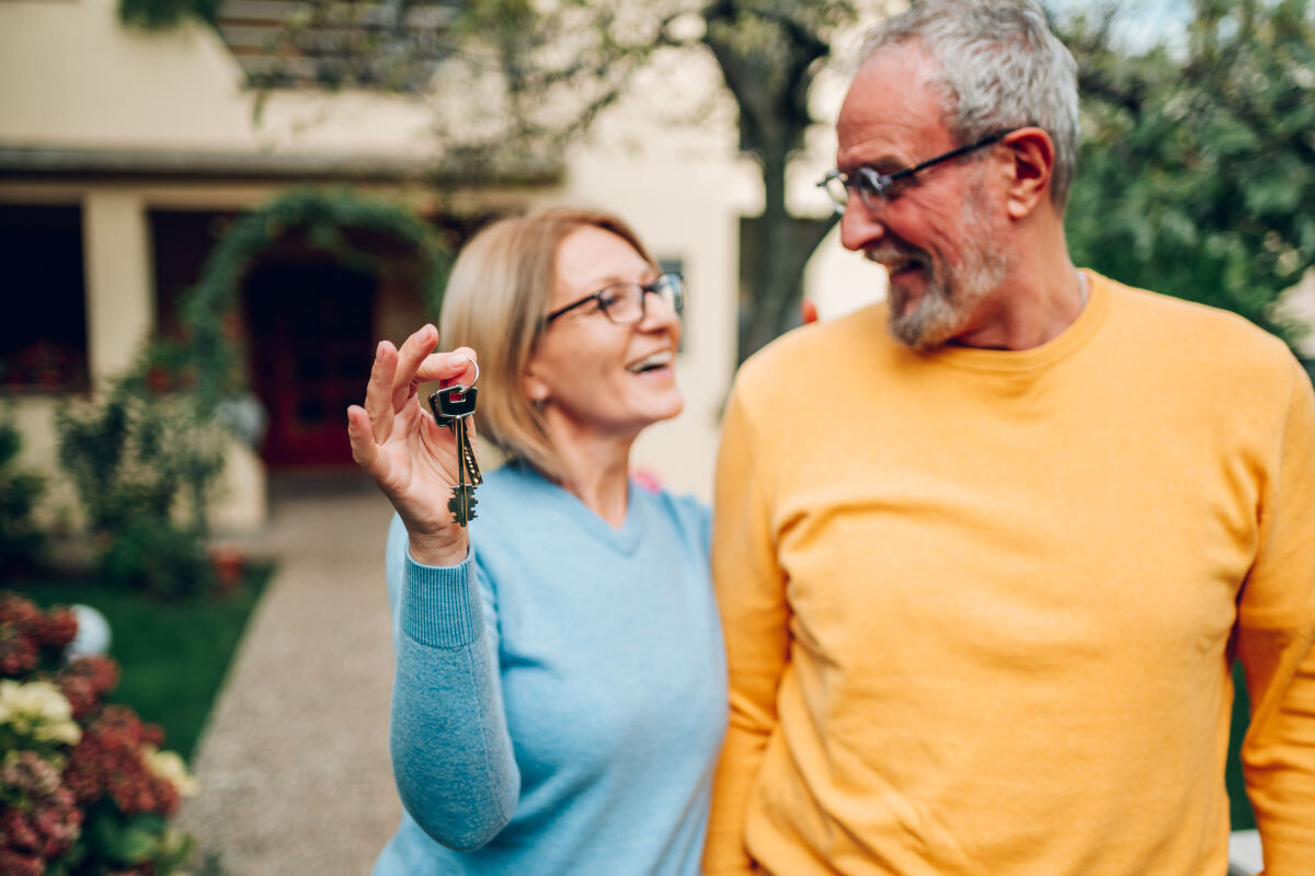 older-couple-holding-keys-standing-in-front-of-home